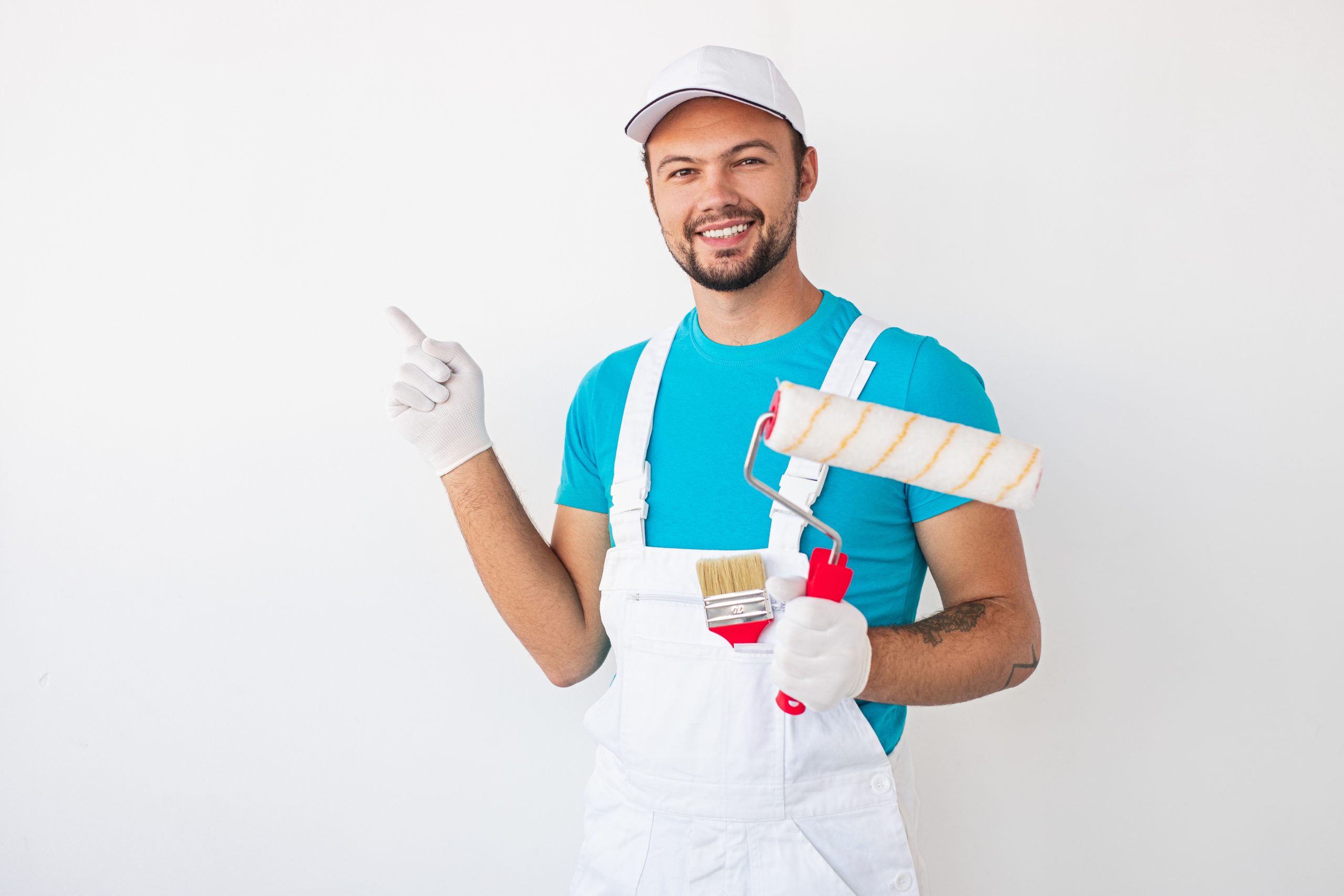 Cheerful young male worker with painting roller and brush pointing at white wall and looking at camera with friendly smile in New York